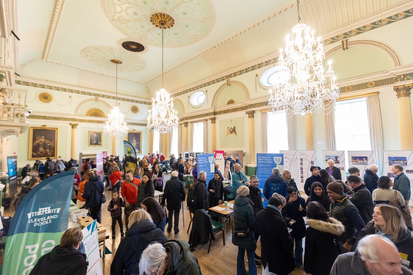 Lots of people amongst display stands and banners in the ornate Banqueting Room at Bath's Guildhall, with large chandeliers hanging above.